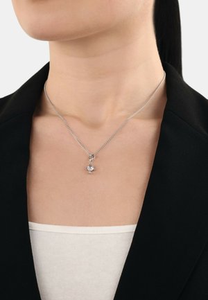Collana d'argento con una delicata catena, dotata di un piccolo ciondolo decorato con una pietra preziosa trasparente, indossata sopra una maglietta bianca e sotto un blazer nero.
