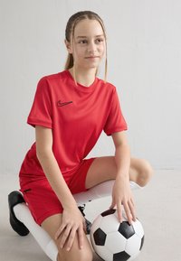 Niña joven con uniforme de fútbol rojo arrodillada con una mano sobre un balón de fútbol blanco y negro frente a un fondo liso.