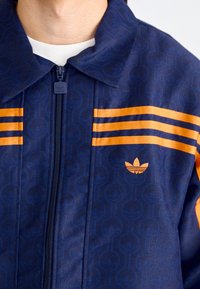 Marinblå zip-up jacka med geometriskt mönster, orange ränder på axlarna och en broderad logotyp på bröstet. Mjuk känsla.