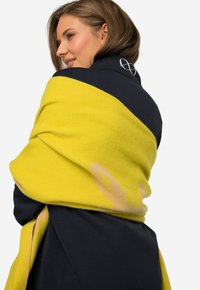 Bufanda amarilla y azul marino, con una textura suave y un acabado liso, decorada con un patrón abstracto. Detalle del logo visible en la zona del hombro.