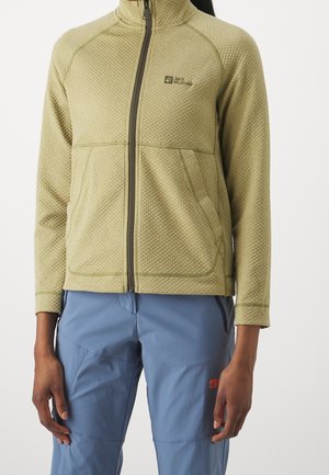 Vrouw die een licht olijfgroene jas met textuur en een ritssluiting draagt, gecombineerd met blauwe outdoorbroek met ritszakken.