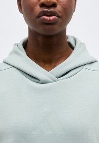 Κοντινό πλάνο του κάτω μέρους του προσώπου και του λαιμού ενός ατόμου που φοράει ανοιχτό γκρι φούτερ με κουκούλα, με αχνό λογότυπο Adidas στο στήθος, πάνω σε απλό φόντο.