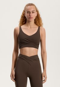 Barna sportmelltartó V-nyakkivágással és széles pántokkal, csavart elülső dizájnnal. Magas derekú barna leggings-szel párosítva, sima anyagból készült.