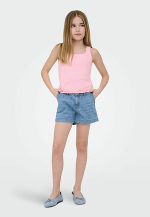 Młoda dziewczyna stojąca z dłońmi w kieszeniach, ubrana w różowy bezrękawnik, niebieskie dżinsowe szorty i jasnoniebieskie baleriny.