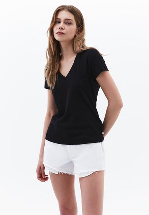 Jonge vrouw met lang blond haar, een zwarte V-hals T-shirt en witte versleten denimshorts, staand voor een effen witte achtergrond.