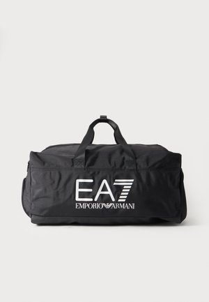 Musta duffel-laukku, jossa kaksi kahvaa ja valkoinen EA7 Emporio Armani -logo edessä, yksinkertaisella vaalealla taustalla.