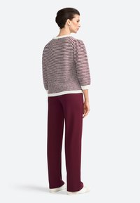 Modèle de dos, portant un pull rayé et un pantalon bordeaux. Des chaussures simples et un fond neutre mettent en valeur la tenue.