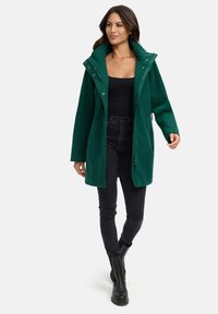Grüner Zip-Up-Mantel mit einer großen Kapuze, Druckknöpfen, Seitentaschen und einem lockeren Schnitt. Getragen über einem schwarzen Oberteil und kombiniert mit schwarzen Jeans und Stiefeln.