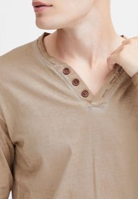 Beige puuvillane pikkade varrukatega särk V-kaelusega, kolme pruuni nupuga peal, pehme ja siledat tekstuuri.
