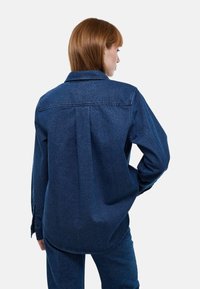 Chemise en denim bleu foncé à coupe décontractée, manches longues, poignets à boutons et empiècement au dos. Présente un ourlet arrondi et un tissu texturé.