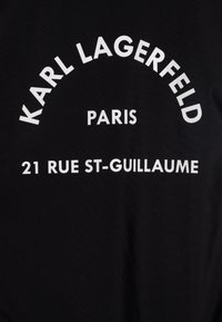 Tecido preto com texto branco em destaque. O texto inclui "KARL LAGERFELD", "PARIS" e "21 RUE ST-GUILLAUME" em tamanhos e curvaturas variadas.