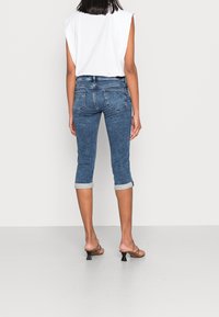 Femme portant un jean en denim bleu raccourci avec revers et un top blanc sans manches, debout sur un sol et un mur gris clair uni.