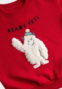 Sudadera roja con un personaje yeti blanco y esponjoso que lleva un gorro de lana azul y blanco. El texto arriba dice "LISTO YETI" en negro.