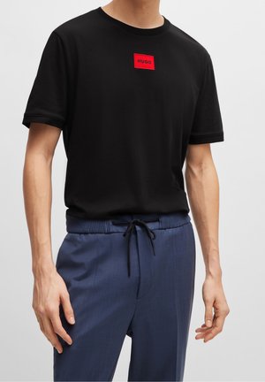 Hombre vistiendo una camiseta negra de manga corta con un parche rojo de logo "HUGO" y pantalones azul con cordón, sobre un fondo plano.