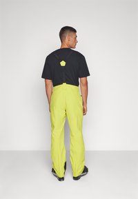 Geel waterdichte broek met verstelbare banden, een gladde textuur en een zwart shirt, gezien van achteren. Bevat hexagonale accentdetails.