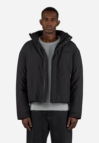 Svart boblejakke med hette, glidelås foran og sidelommer. Har en teksturert grå genser under og mørke jeans. Enkel, moderne design.