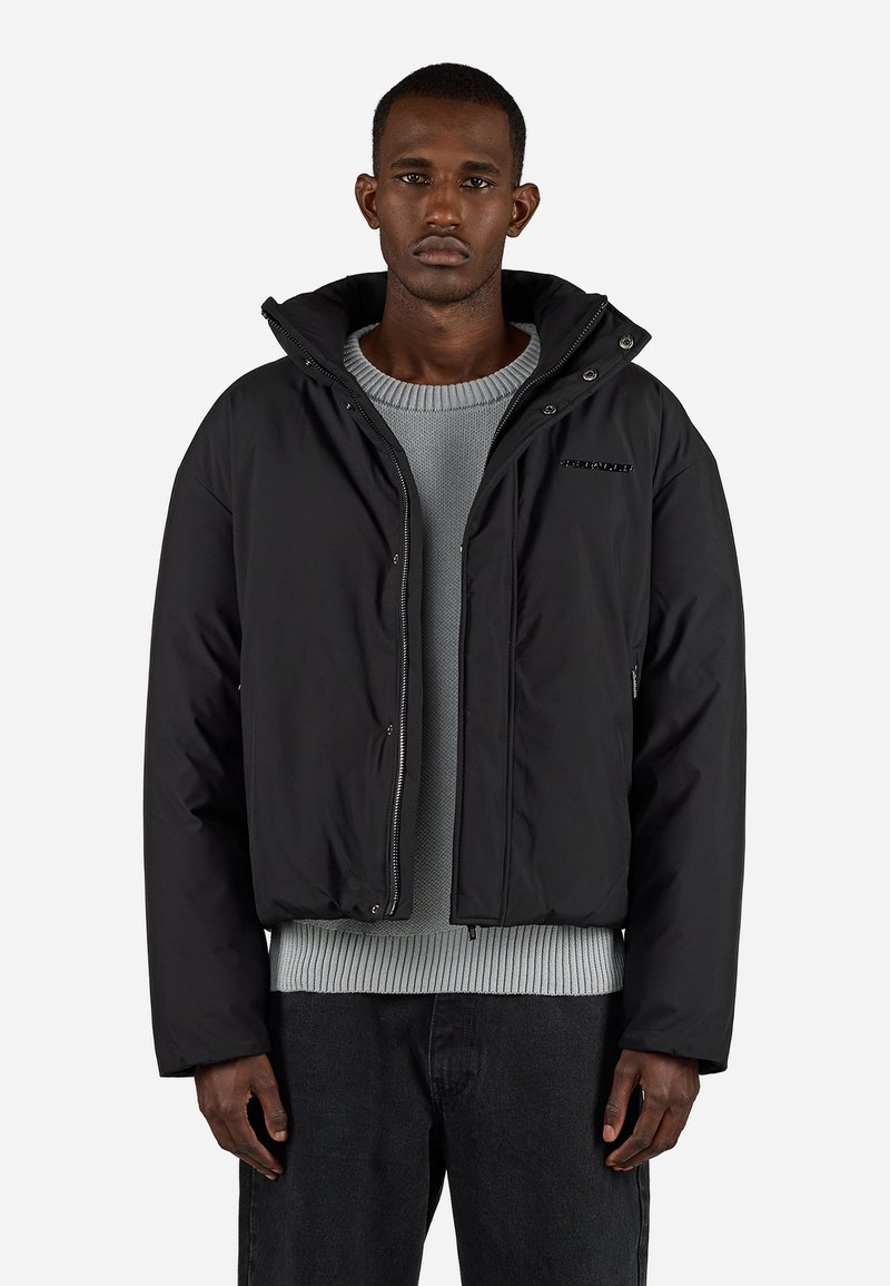Svart boblejakke med hette, glidelås foran og sidelommer. Har en teksturert grå genser under og mørke jeans. Enkel, moderne design.