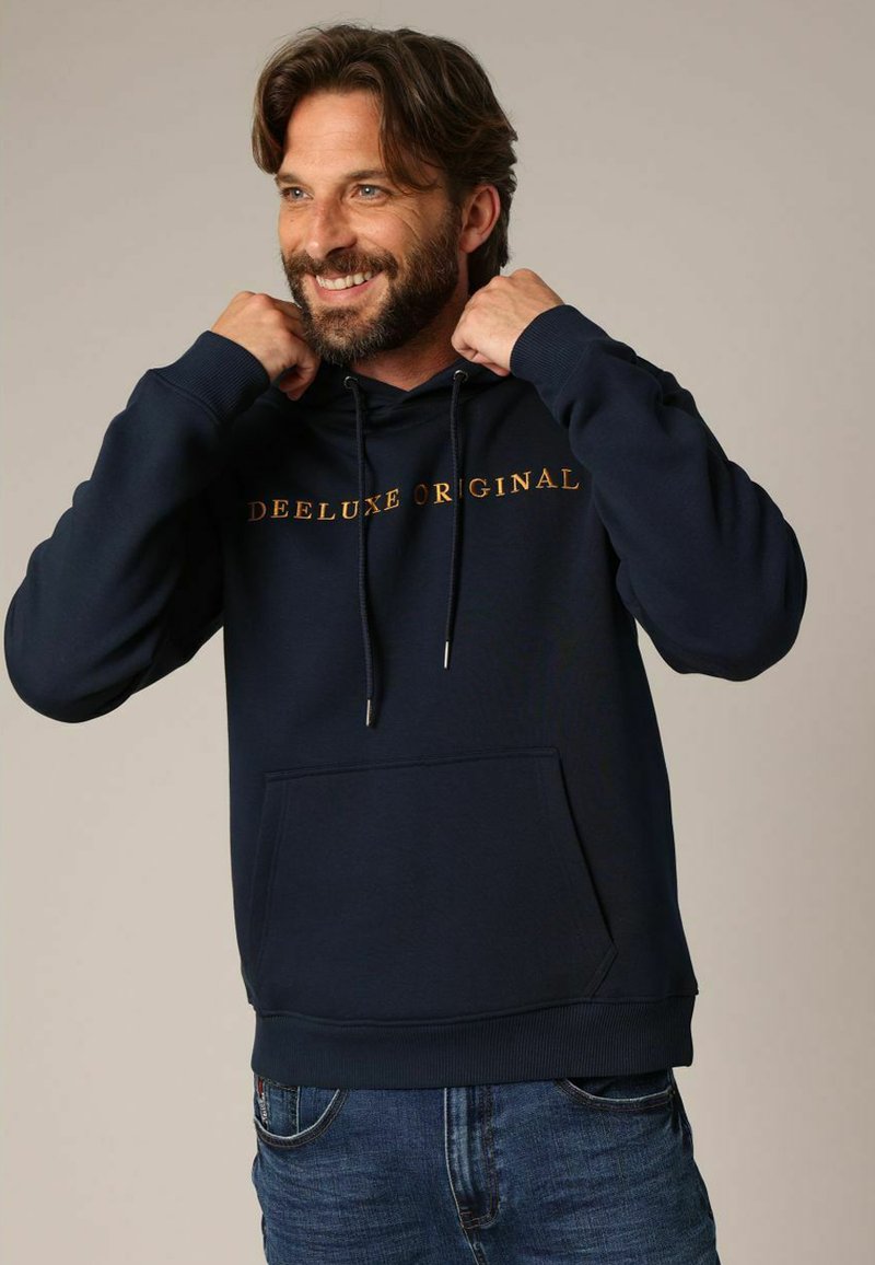 Hoodie bleu marine en tissu doux, doté d'une poche kangourou, d'une capuche avec cordon de serrage réglable, et de "DEELUXE ORIGINAL" en texte doré contrastant.