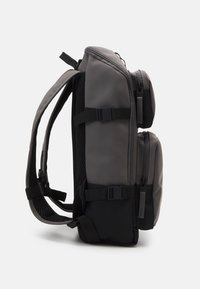 Grauer Rucksack mit sleekem Design, zwei Seitenfächern, verstellbaren Trägern, Mesh-Details und robusten Reißverschlüssen. Hergestellt aus wasserabweisendem Material.