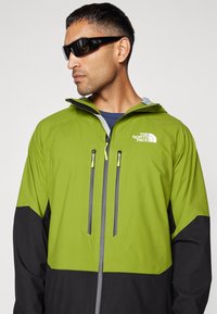 Zielona i czarna wodoodporna kurtka z kapturem, wyposażona w kieszenie zapinane na zamek, z logo na klatce piersiowej oraz kontrastowymi szarymi akcentami.