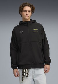 Chaqueta negra ligera con capucha, con cierre frontal de cremallera, puños elásticos y logotipos de Aramco y Puma. Incluye un detalle de lazo a cuadros en el dobladillo.