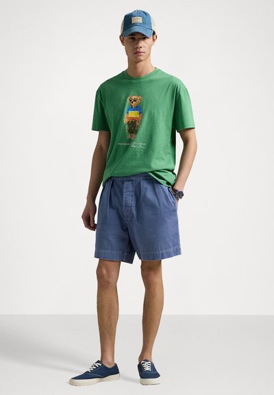 T-shirt en coton vert avec un motif d'ours, accentué de bleu et jaune. Shorts bleu clair avec une taille élastique. Chaussures bleues avec des lacets blancs.