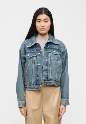 Jeune femme aux longs cheveux foncés, portant une veste en jean bleue cloutée par-dessus un pull gris et un pantalon beige, posant devant un fond uni.