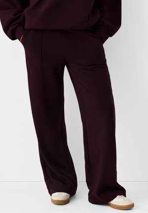 Pantalon large couleur violet foncé porté avec des baskets blanches, mains dans les poches, debout devant un fond clair uni.