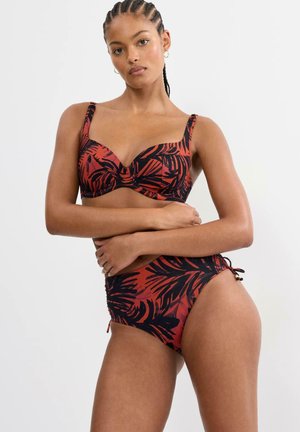 Frau mit geflochtenem Haar trägt rot-schwarzes Bikini mit tropischem Blattmuster, steht mit verschränkten Armen vor hellem Hintergrund.