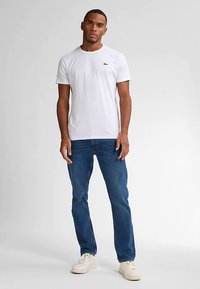 Tricou alb din bumbac cu guler rotund și logo mic pe pieptul stâng, asortat cu blugi denim albaștri și adidași de culoare deschisă.