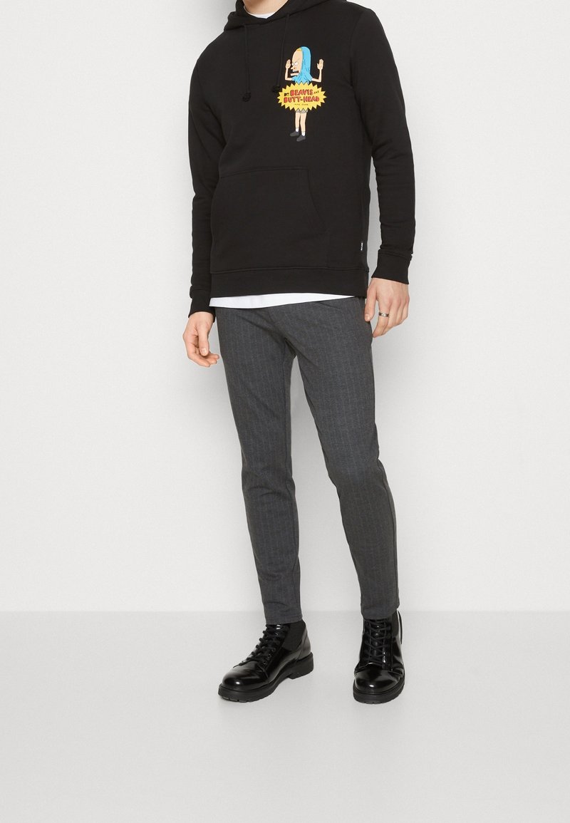 Personne portant un sweat à capuche noir avec un personnage de dessin animé et le texte « Beavis and Butt-Head », pantalon gris slim-fit et bottines noires à lacets, debout devant un fond blanc.