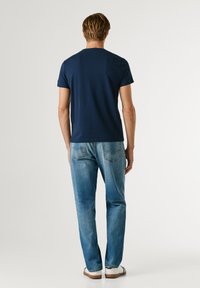 Muž stojaci chrbtom ku kamere, oblečený v tmavomodrom tričku, svetlomodrých džínsach a bielych teniskách na jednofarebnom svetlom pozadí.