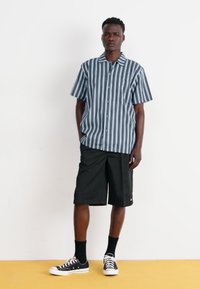 Cămașă cu mâneci scurte în dungi albastre și albe, combinată cu pantaloni scurți negri până la genunchi, șosete negre și adidași negri. Design simplu cu nasturi.