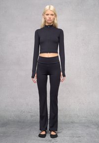 Schwarzes, langärmliges, kurzes Oberteil mit hohem Kragen und Reißverschluss vorne, kombiniert mit hochtaillierten, schwarzen, ausgestellten Leggings und minimalistischen, schwarzen Schuhen.