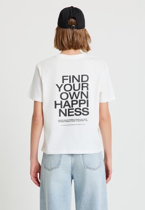 Persona con una maglietta bianca con la scritta "TROVA LA TUA FELICITÀ" sul retro, jeans azzurri e un cappellino nero, vista da dietro.