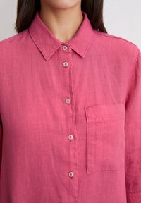 Rozā lina krekls ar apkakli, ar pogu aizdari priekšpusē, piecām pogām ar kontrastējošu šuvumu un krūšpocketu kreisajā pusē.