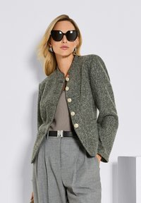 Blonde vrouw met zwarte oversized zonnebril, zilveren ronde oorbellen, grijze getextureerde jas met gouden knopen, en grijze high-waist broek met riem.