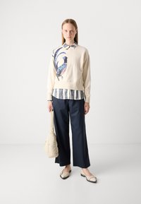 Sudadera crema con gráfico de un pájaro azul, superpuesta sobre una camisa a rayas azul y blanca. Pantalones de pierna ancha en color navy, zapatos planos beige, y un bolso crema.