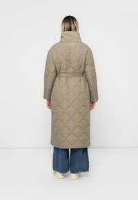 Persoon in een lange beige gewatteerde jas met riem, wijde blauwe jeans en witte sneakers, staand met het gezicht naar een witte muur.