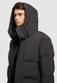 Chaqueta de plumón negra aislante con un alto cuello, gran capucha y parche en el hombro. Presenta una textura suave y cierre de cremallera.