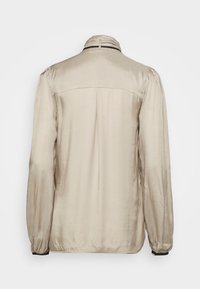 Giacca in satin beige con una texture liscia, maniche lunghe, colletto alto e polsini elasticizzati. Presenta un design semplice senza hardware visibile.