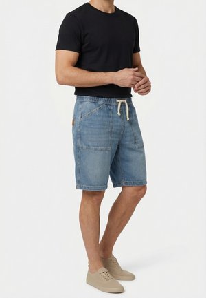 Uomo che indossa una t-shirt nera, pantaloncini di jeans azzurri con coulisse e grandi tasche, e sneakers beige, in piedi davanti a uno sfondo bianco.