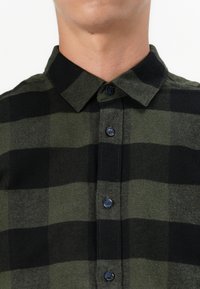 Man wearing green and black checkered button-up shirt, collar buttoned, neck and chin visible, neutral background.