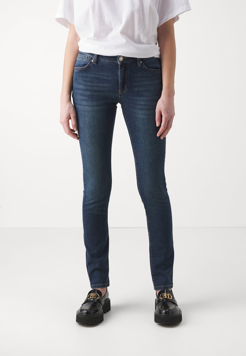 Sötét mosású skinny farmernadrág, közepes derékkal, elülső zsebekkel és diszkrét kopásokkal, fekete loaferekkel párosítva.