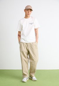 T-shirt bianco a maniche corte con logo, pantaloni beige larghi e sneakers bianche con dettagli verdi. Cappellino color sabbia indossato sulla testa.