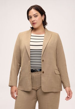 Donna con capelli corti e scuri che indossa un completo beige e una maglia a righe bianca e nera, in piedi contro uno sfondo chiaro e semplice.
