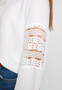 Blouse blanche à manches avec une incrustation en dentelle florale et des rayures horizontales, portée avec un jean en denim bleu, sur un fond gris clair.