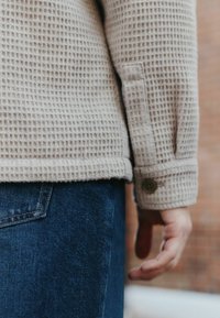 Beige textured sweater with a waffle weave design and buttoned cuffs, paired with dark blue denim jeans. Close-up of the sleeve and hand.
