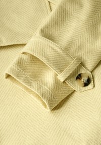 Světle žlutá látka bundy s herringbone vzorem, s ohnutým manžetou a kulatým knoflíkem s mramorovým designem.