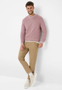 Lichtroze textuur gebreid sweater met ronde hals, beige broek en beige sneakers met witte zolen. Model staat tegen een witte achtergrond.